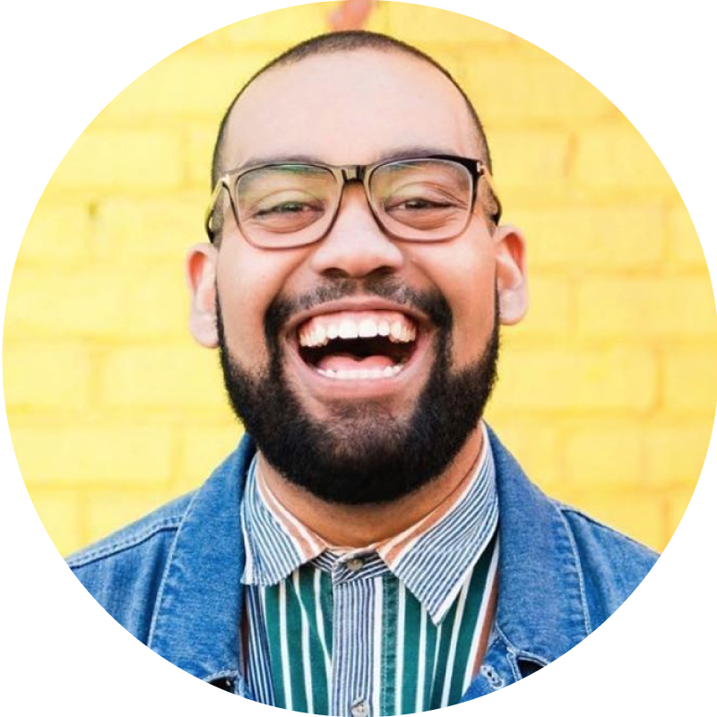 Photo of Ben O'Keefe, a young Black man with a beard facing forward with a big smile, wearing glasses and jean jacket over a striped collared shirt in front of a yellow background. 